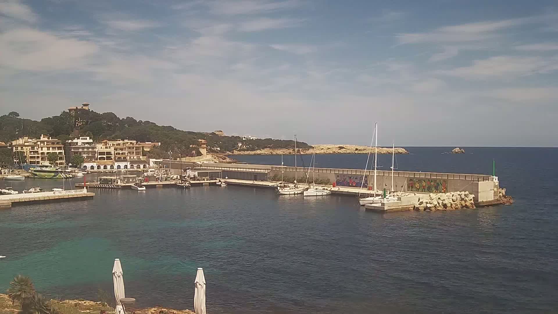 A small harbor filled with boats is nestled next to a town of low-rise buildings on a sunny day with partly cloudy skies.