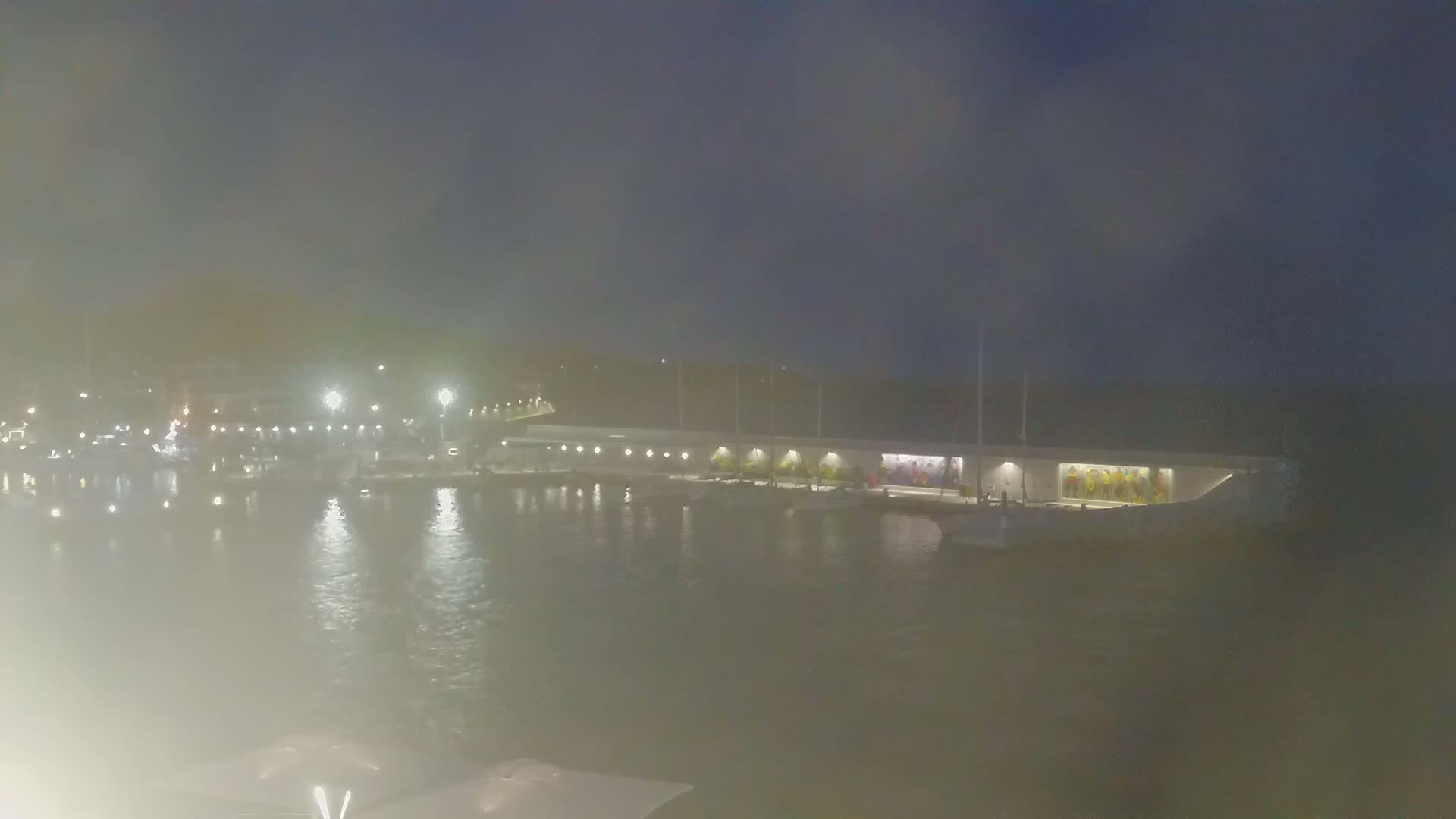 A hazy night scene shows a marina with boats docked along a long building illuminated by lights.