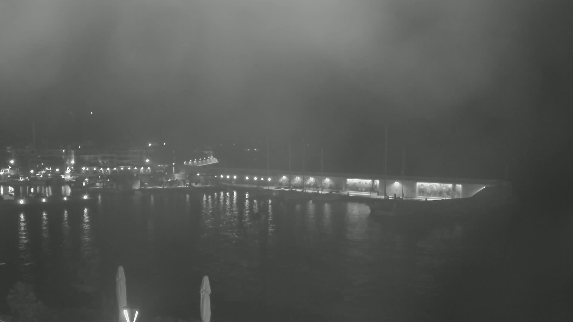 A foggy night scene shows a harbor with boats moored along a lit waterfront, under a dark sky.