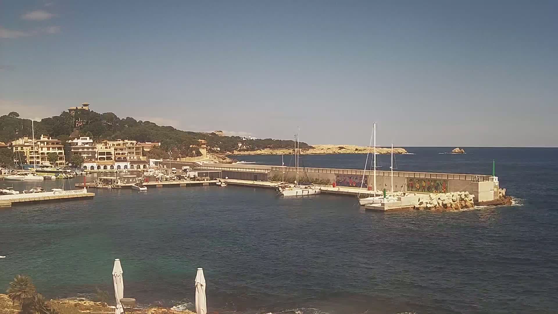 A small harbor with docked sailboats is situated near a town nestled against a hillside under a clear, sunny sky.
