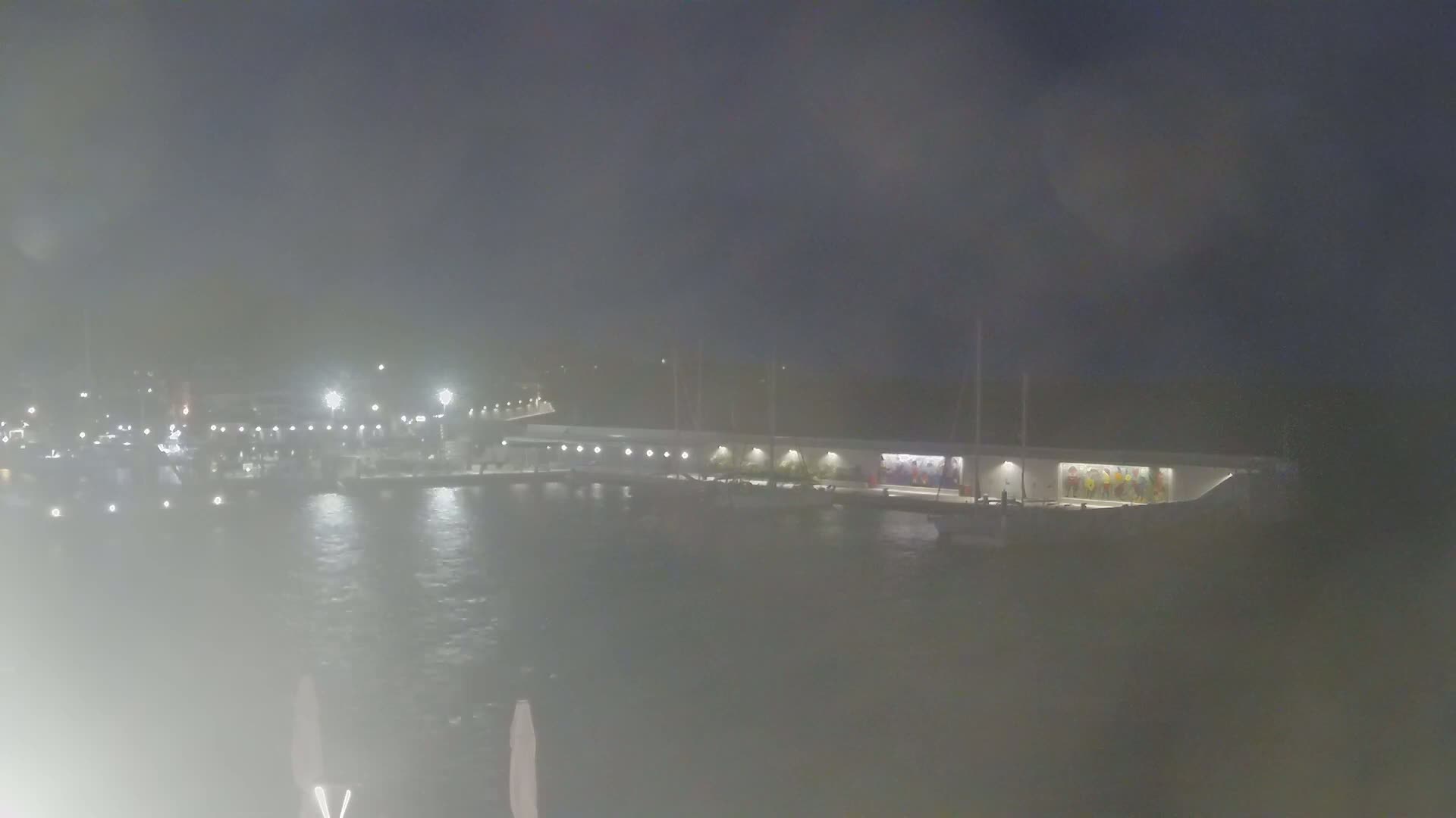 A hazy night scene shows a marina with docked boats and a long building with colorful artwork on its side, illuminated by lights.