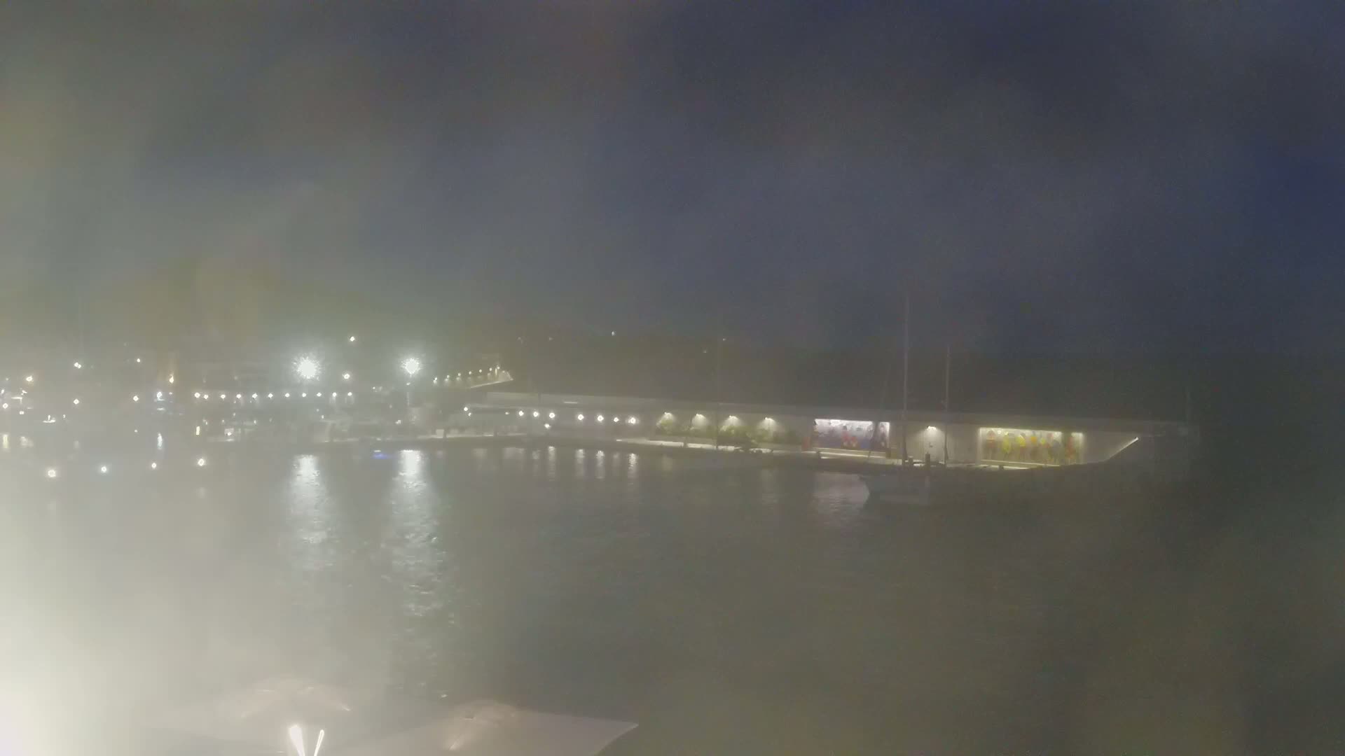 A hazy nighttime view shows a marina with boats moored along a waterfront building illuminated by lights.
