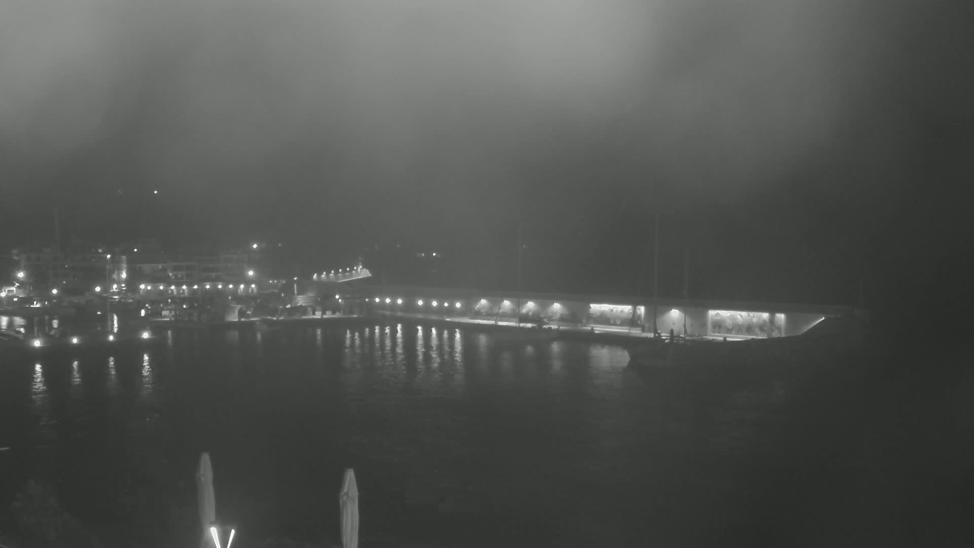 A foggy nighttime scene shows a waterfront area with buildings and lights reflecting on calm water.