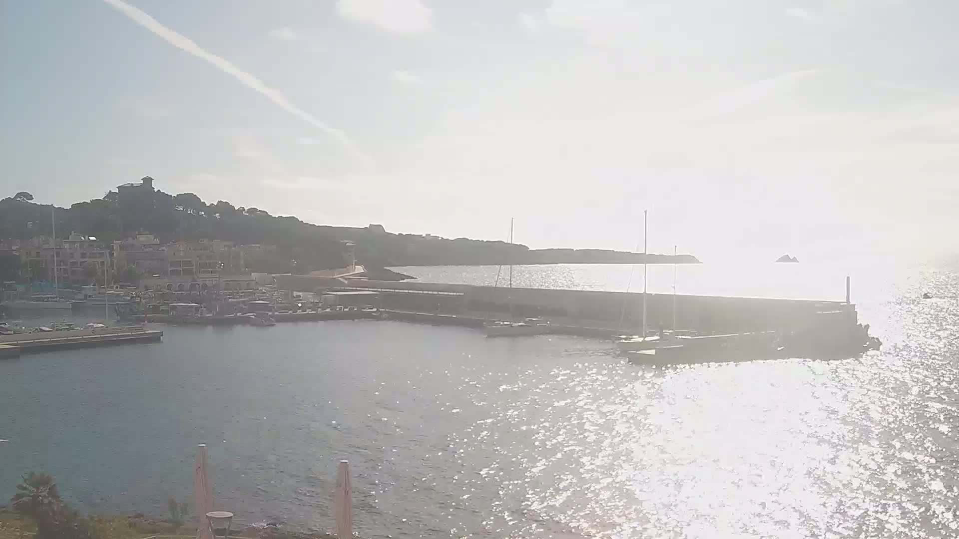 A marina filled with boats is situated beside a calm, sunlit sea, with hills and buildings visible in the background under a mostly clear sky.