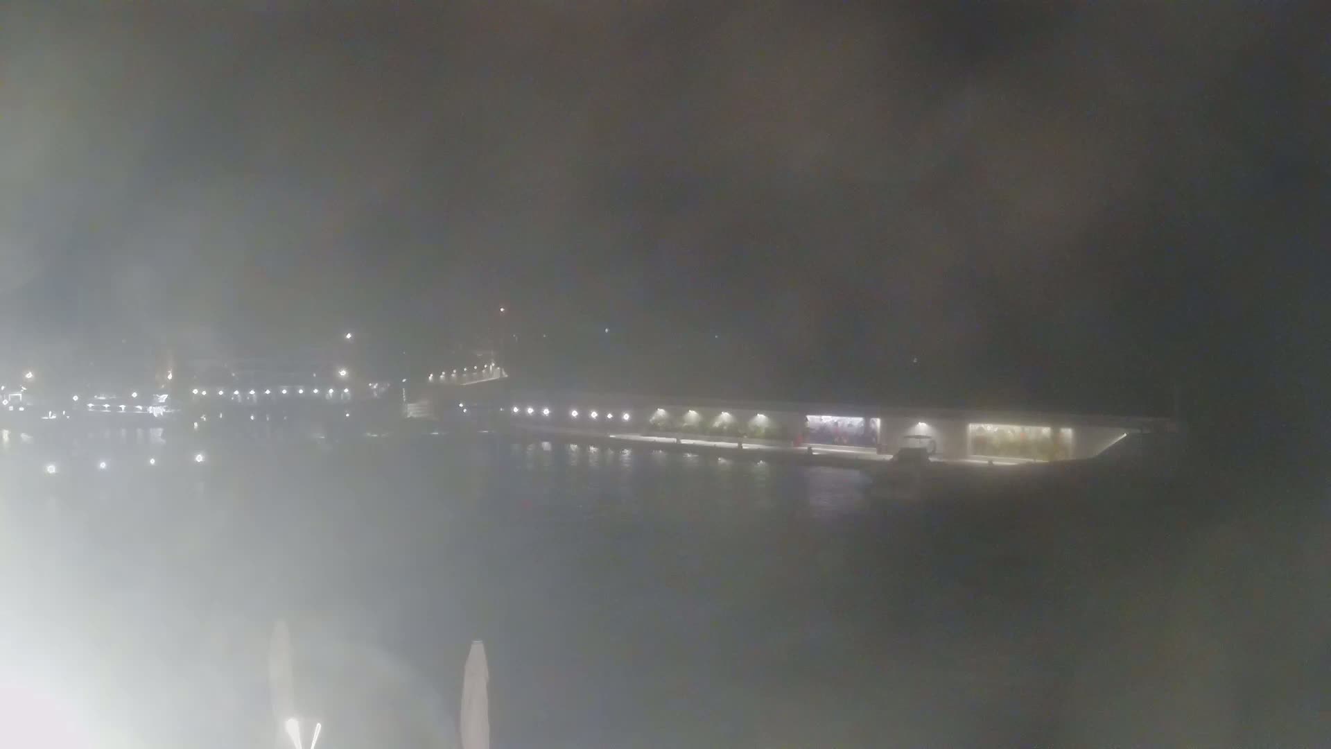 A foggy nighttime scene shows a long, low building alongside a body of water, with lights visible on the building and across the water.
