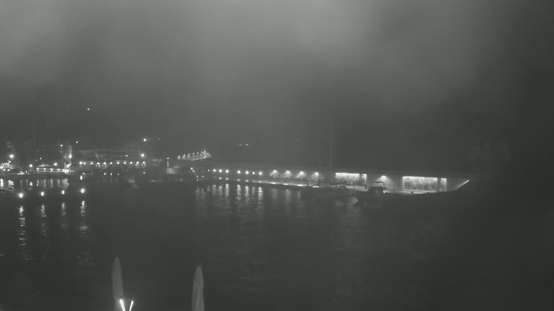 A foggy nighttime scene shows a body of water with lights reflecting on its surface, a waterfront area with buildings and boats, and a thick fog obscuring the background.