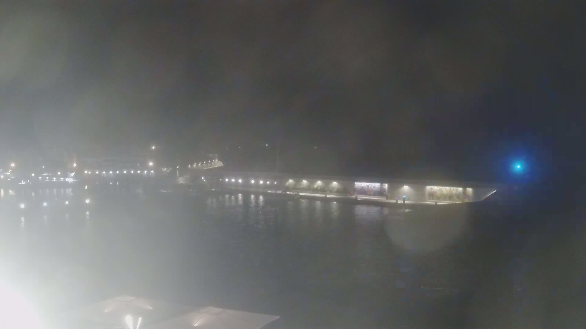 A hazy night scene shows a long, low building lit by several lights along the water's edge.