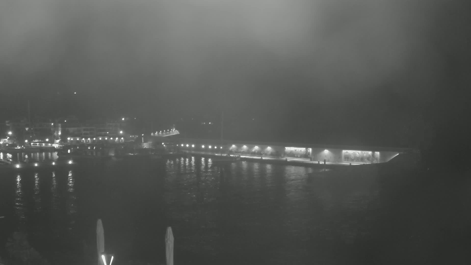 A foggy nighttime scene shows a waterfront with boats in a marina and a long, low building illuminated along a calm body of water.