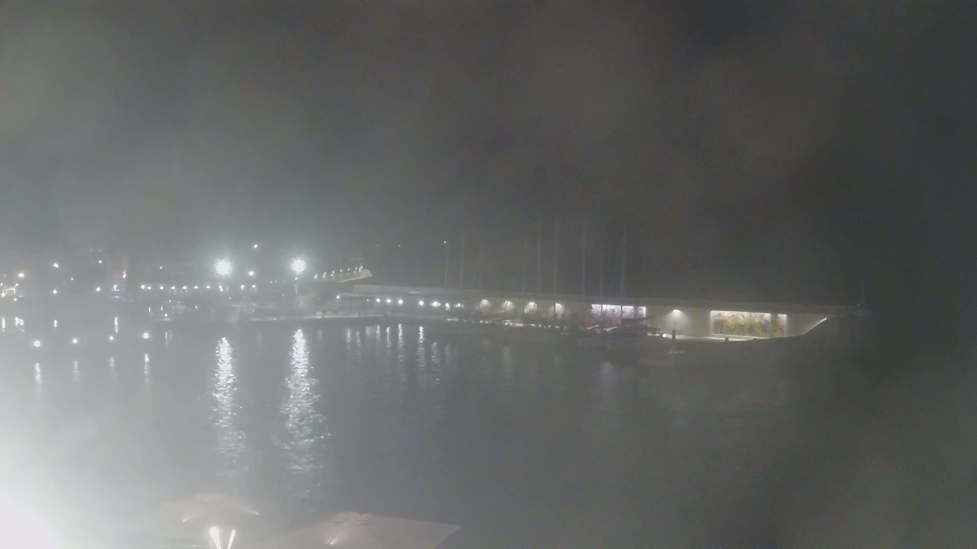 A hazy night scene shows a waterfront building with lights reflecting on calm water.