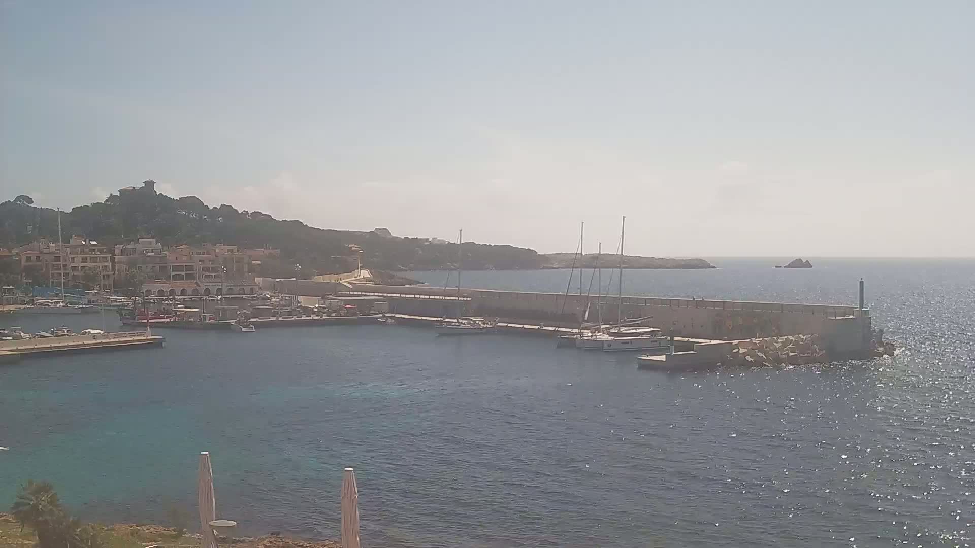 A calm, sunny day reveals a harbor filled with sailboats, nestled beside a hillside town and a long stone wall extending into the sea.