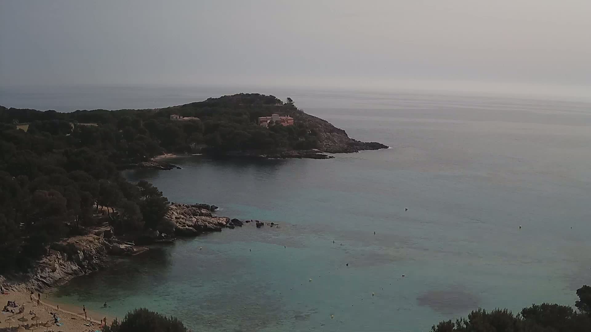 A hazy day reveals a secluded cove with a small sandy beach and calm, clear turquoise water, surrounded by dark green vegetation and rocky terrain.