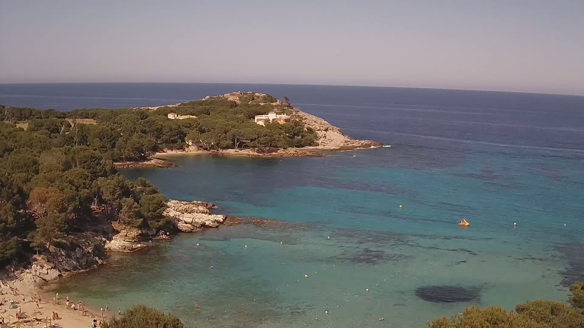 A secluded cove with a sandy beach and clear turquoise water is surrounded by lush green vegetation and a rocky headland, under a bright sunny sky.