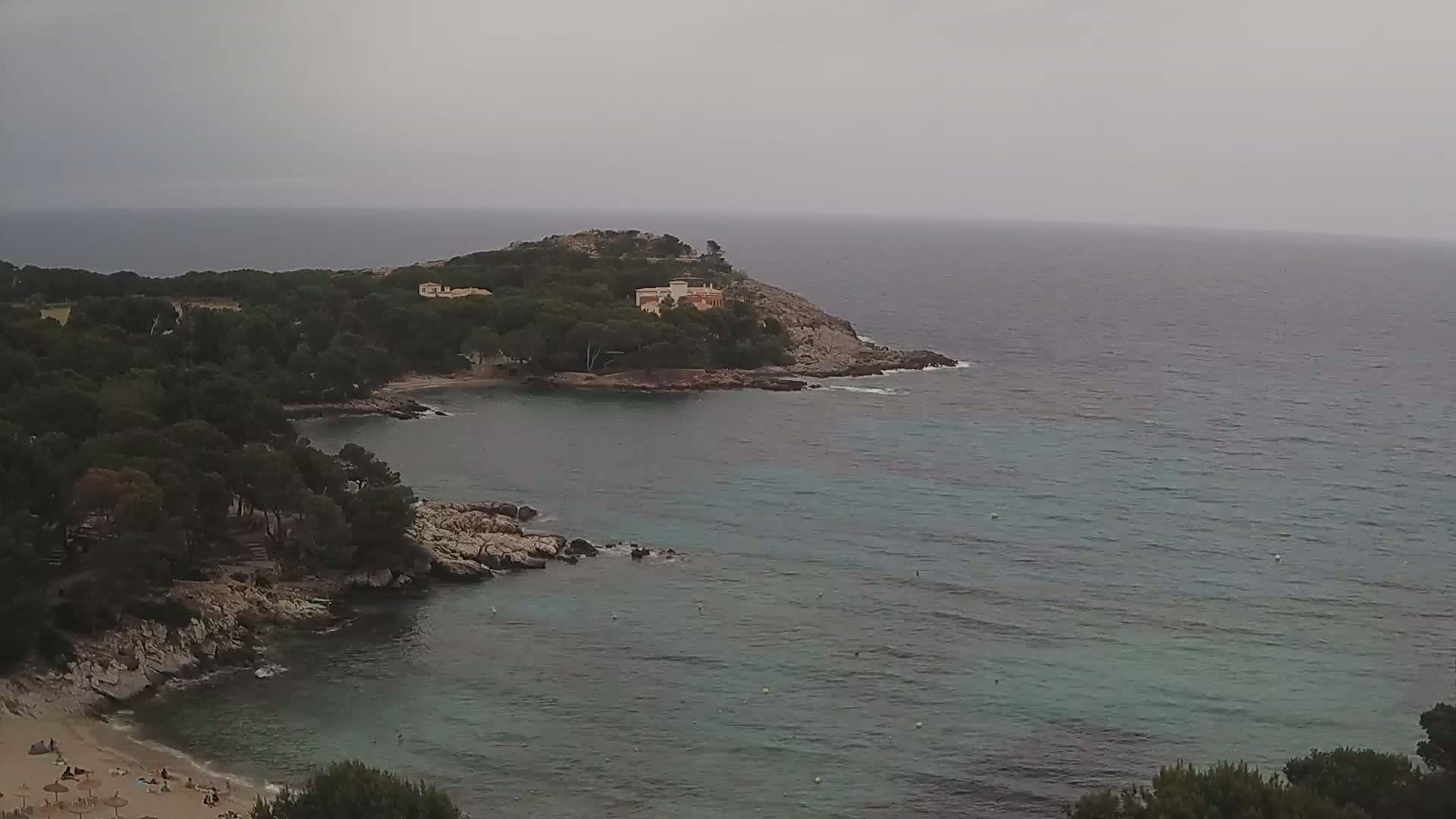 A secluded cove with a small beach, surrounded by dark green trees and rocky coastlines, opens to a calm, gray-green sea under an overcast sky.