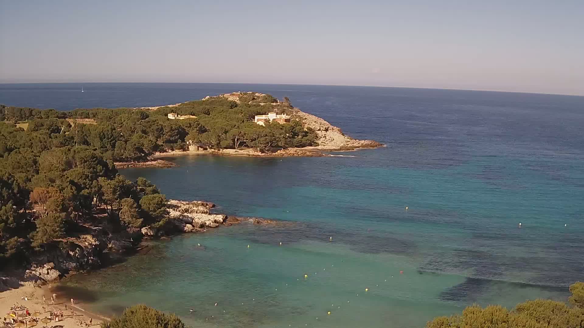 A secluded cove with a sandy beach and turquoise water is surrounded by pine trees and a small building on a rocky point, under a clear sunny sky.