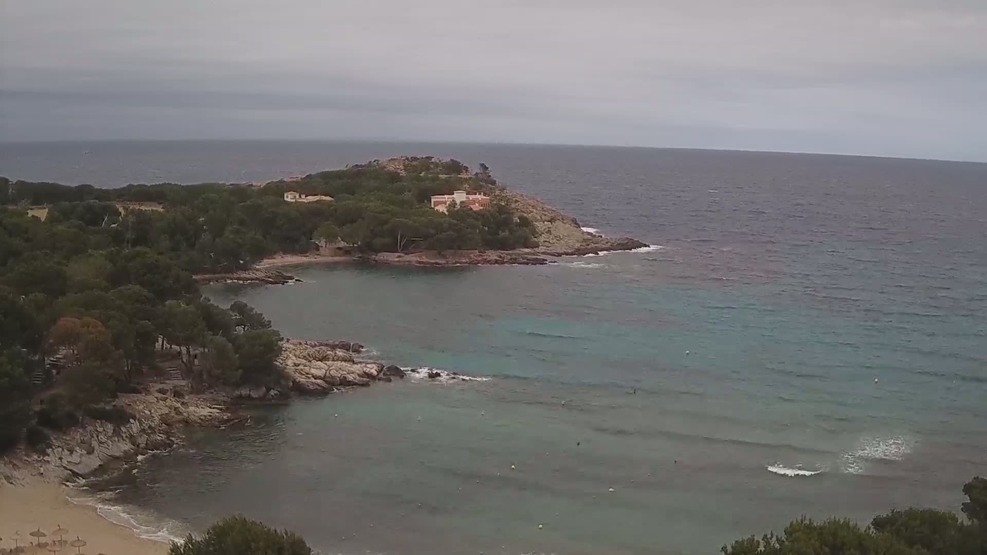 A tranquil cove with a sandy beach, surrounded by pine trees and rocky outcrops, meets a calm, teal sea under an overcast sky.
