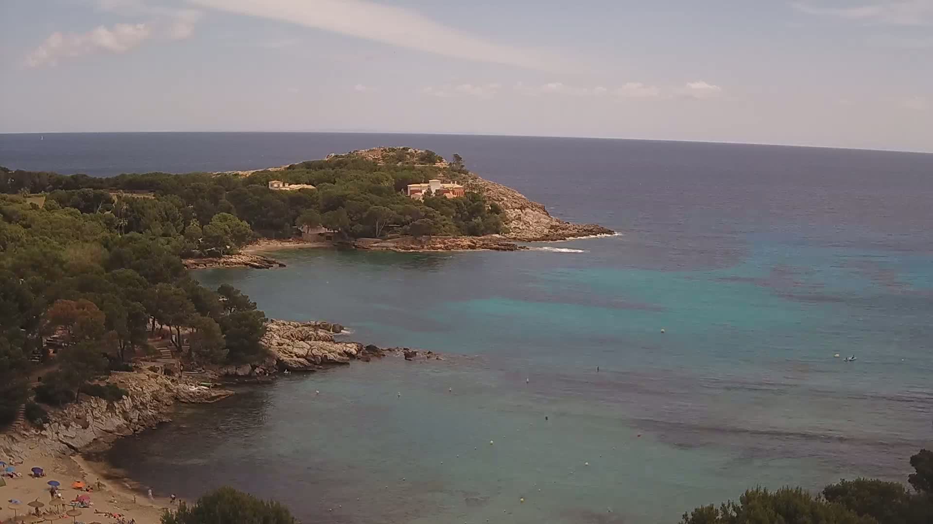 A secluded cove with a sandy beach and turquoise water is surrounded by rocky shores and lush green trees under a partly cloudy sky.