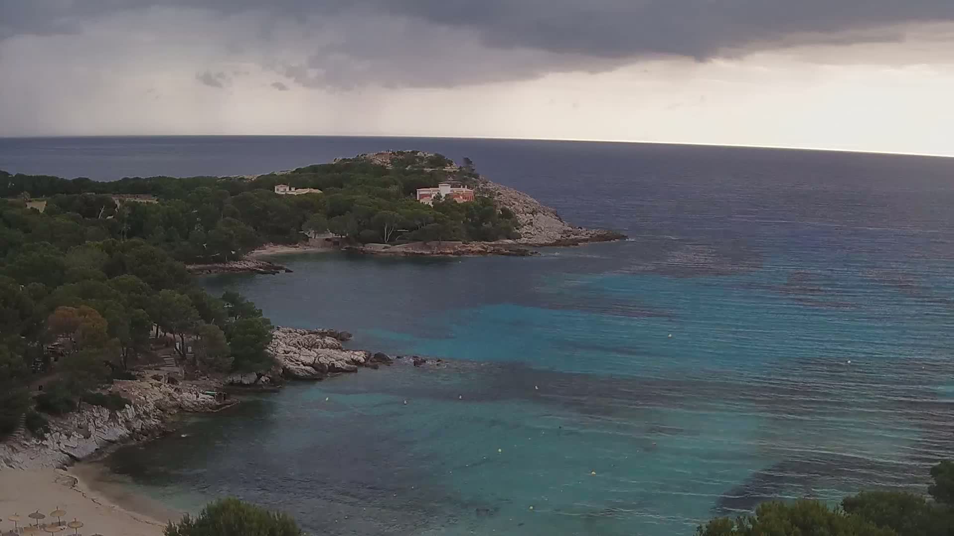 A secluded cove with a sandy beach, surrounded by dark green trees and rocky shores, is partially obscured by a dark, stormy sky over a turquoise sea.