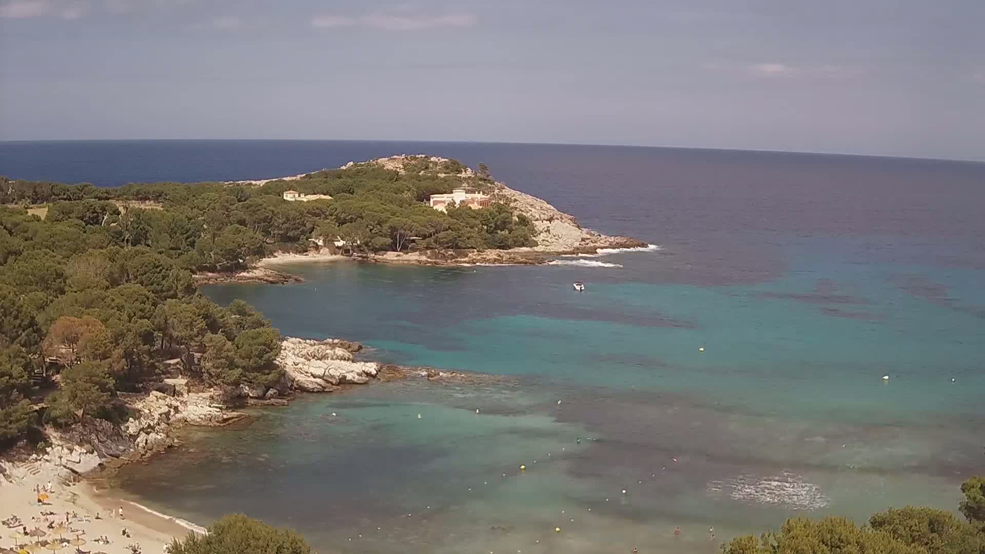 A secluded cove with clear turquoise water, a sandy beach with people, and a rocky headland topped with trees and a building is visible under a mostly sunny sky.