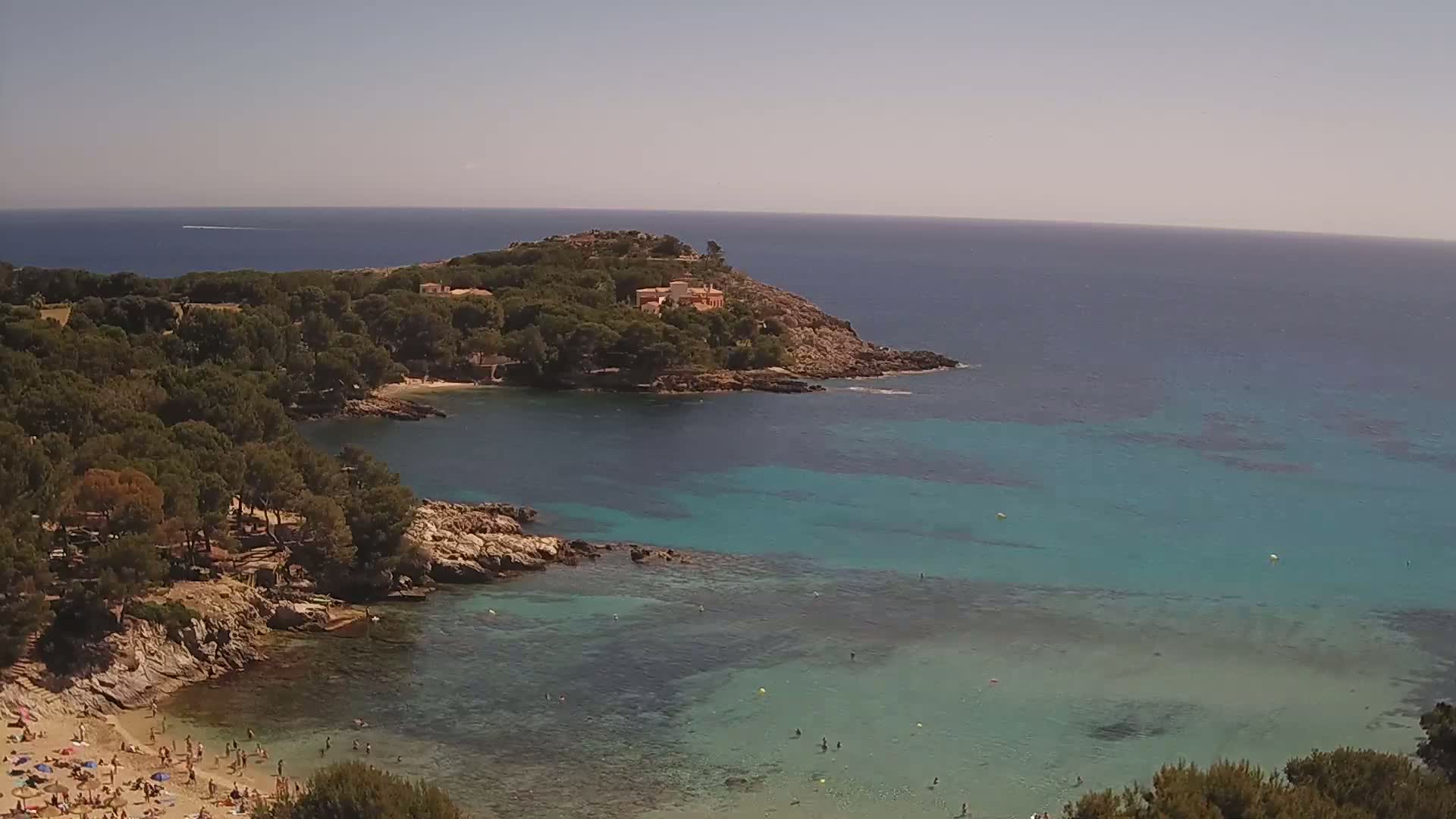 A sunny day reveals a secluded cove with clear turquoise water, a sandy beach with people swimming and sunbathing, and a rocky headland covered with lush green trees.