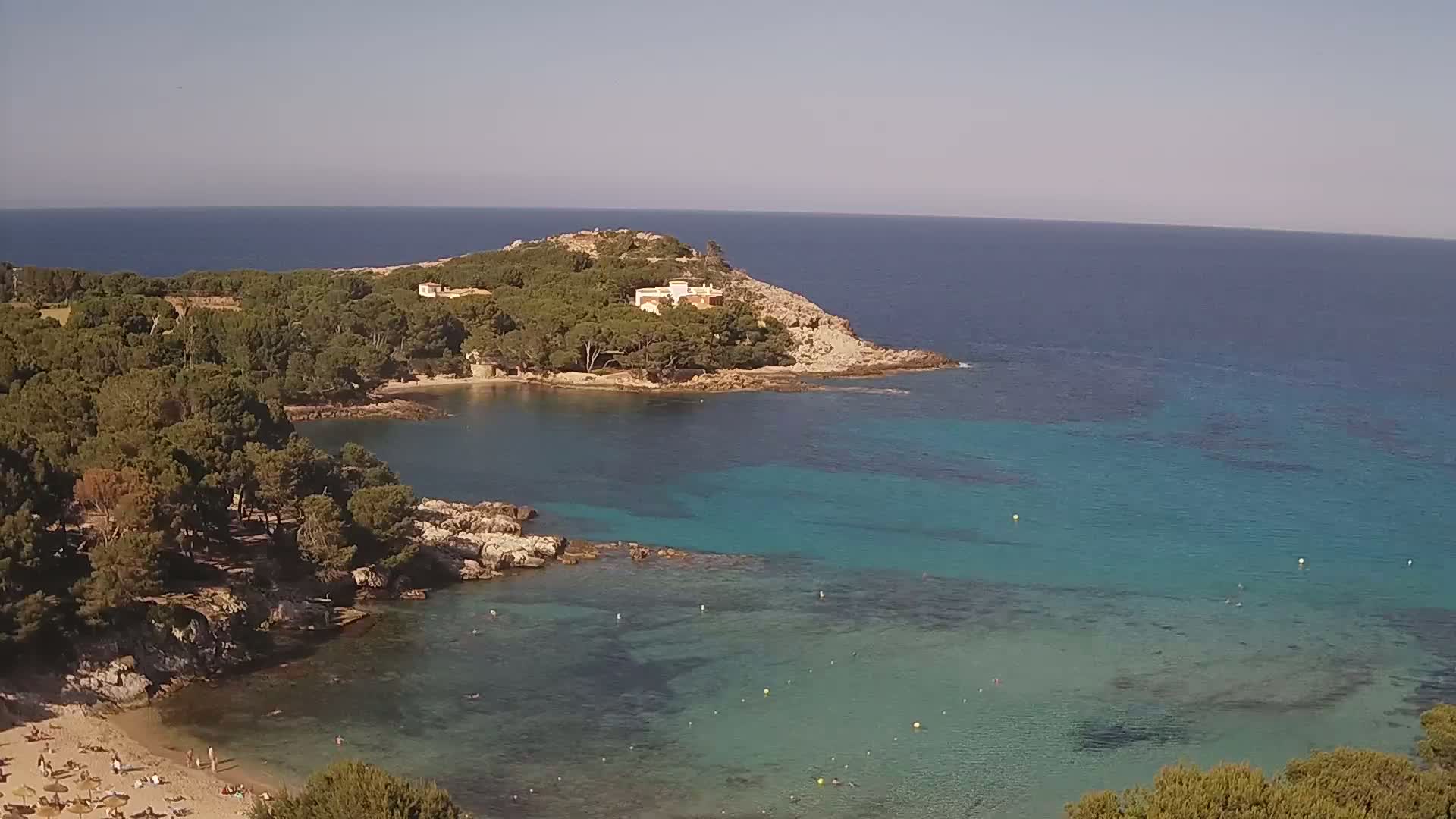 A secluded cove with a sandy beach and clear turquoise water is surrounded by pine trees and a small house on a rocky headland under a sunny sky.