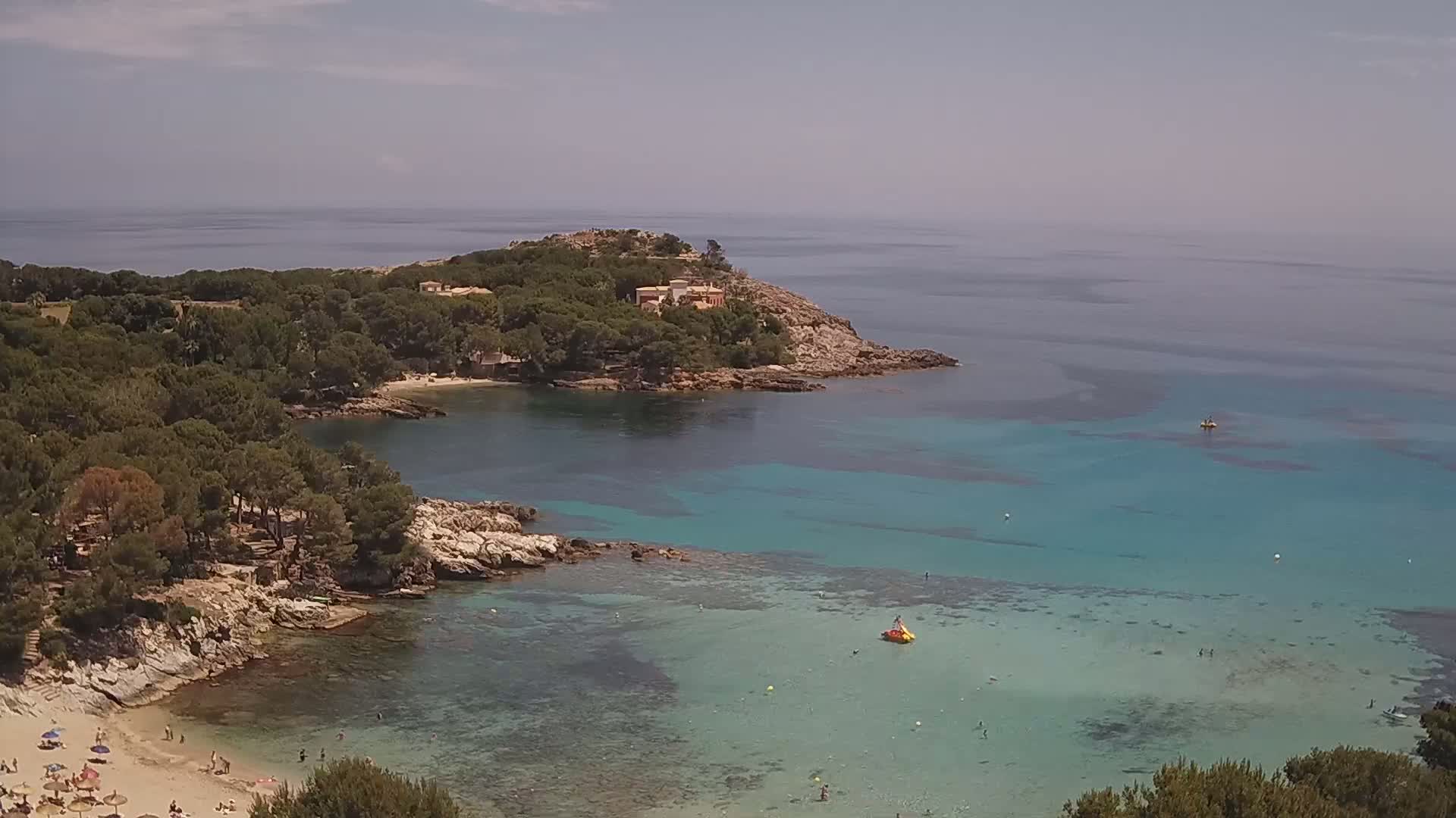 A rocky cove with a sandy beach and clear, turquoise water is surrounded by pine trees under a partly cloudy sky.