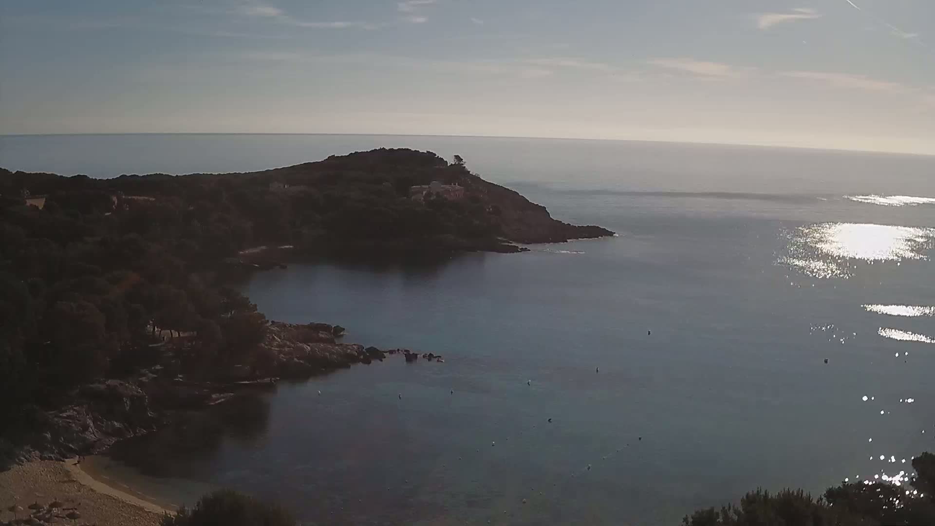 A tranquil cove with a small sandy beach is nestled amongst dark green vegetation, overlooking a calm sea reflecting sunlight under a mostly clear sky.