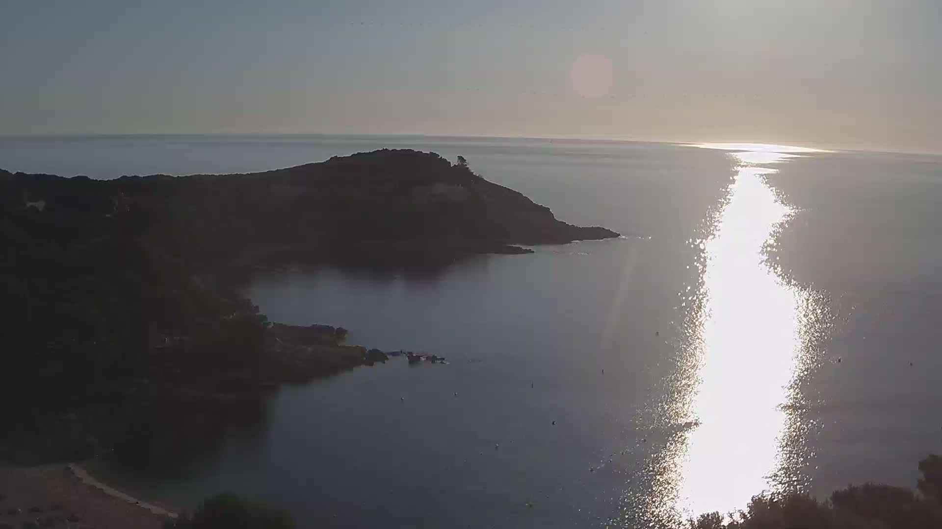 A tranquil coastal scene under a bright, sunny sky shows a calm sea reflecting sunlight, with dark green hills and a small peninsula extending into the water.