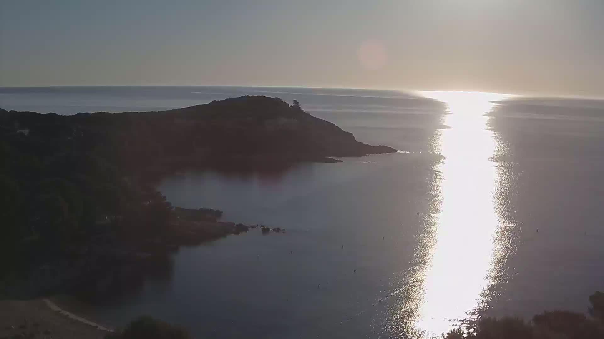 A tranquil coastal scene at sunset shows a dark, rocky peninsula jutting into a calm sea where the sun's reflection creates a bright path across the water.