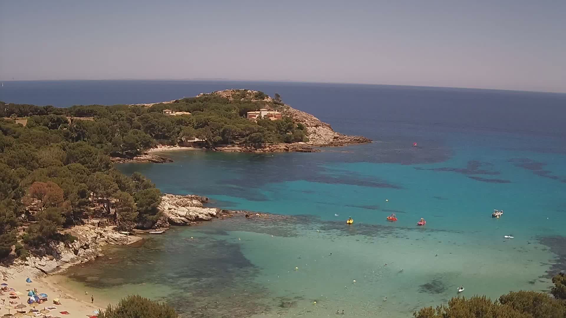 An aerial view shows a sandy beach with sunbathers, clear turquoise water with several small boats, and a rocky coastline covered with lush green trees under a bright sunny sky.