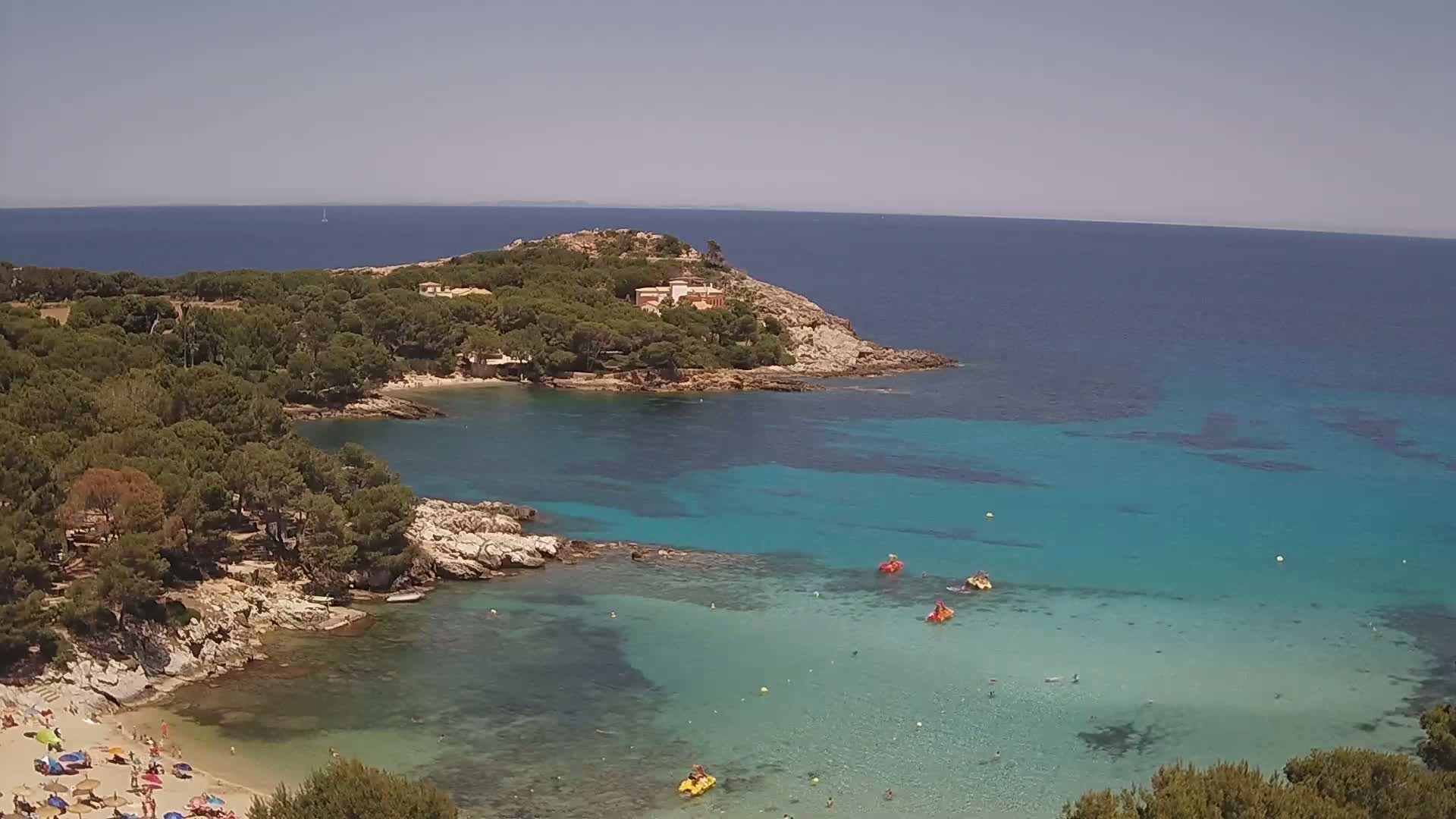 An aerial view shows a secluded cove with clear turquoise water, a sandy beach with people, and a rocky headland covered in lush green trees under a clear, sunny sky.