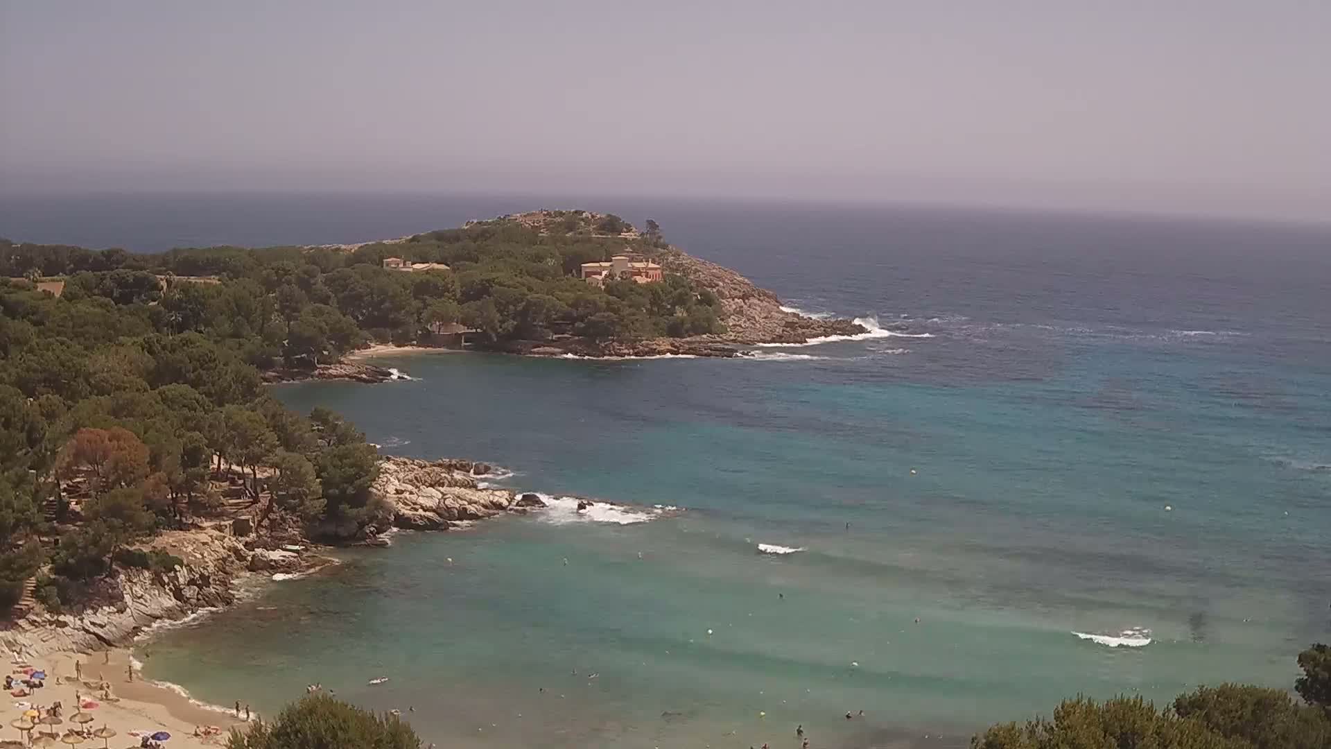 A hazy, sunny day reveals a secluded cove with a sandy beach and turquoise water, surrounded by lush green trees and rocky shores, culminating in a small headland with buildings.