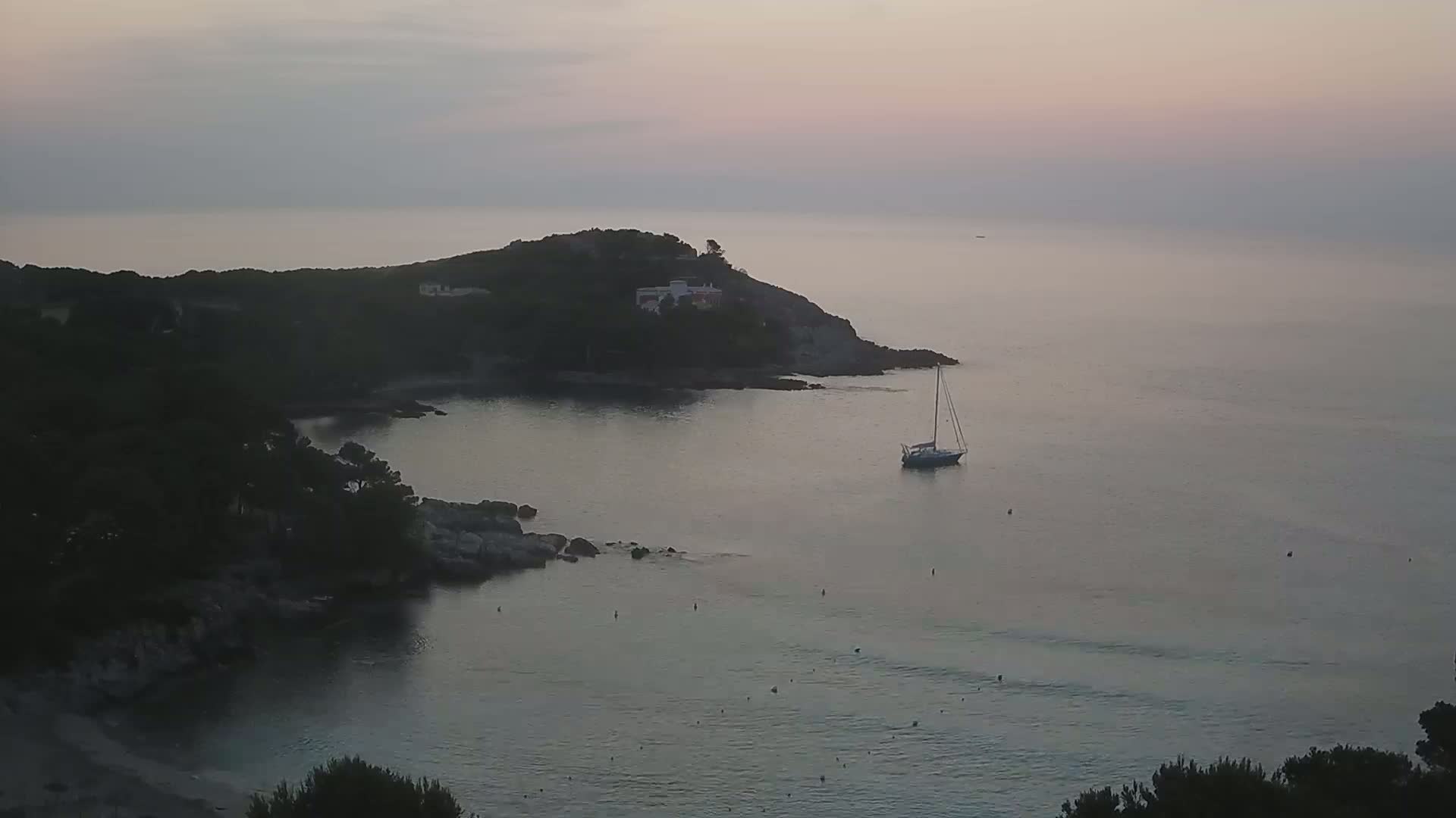 A sailboat is anchored in a calm, dusky-lit bay, nestled beside a small, tree-covered headland with a house visible on its top.