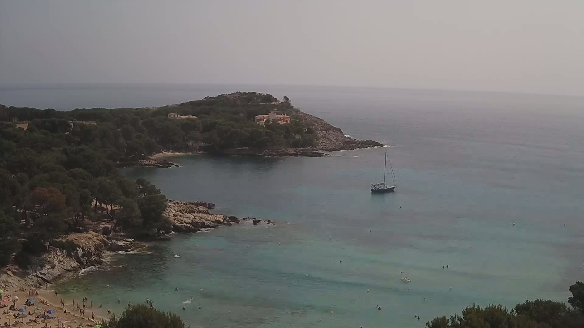 A hazy day shows a secluded cove with a sandy beach and several people swimming and sunbathing, a sailboat anchored offshore, and a small residential area on a rocky headland.