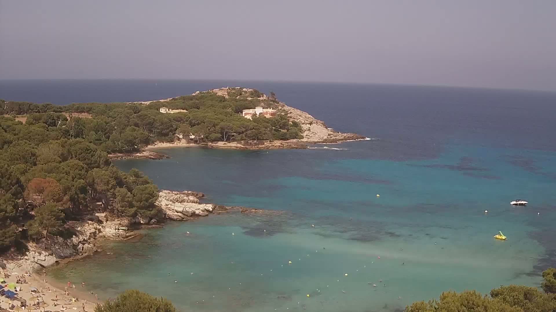 A secluded cove with a sandy beach and clear turquoise water is surrounded by pine trees and rocky terrain under a hazy sky.