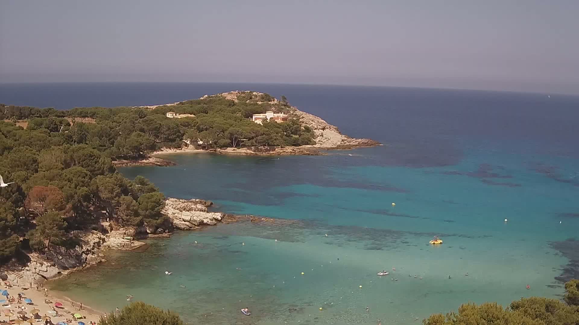 A sandy beach with sunbathers is nestled in a cove of clear, turquoise water surrounded by a rocky shoreline and pine-covered hills under a bright, sunny sky.