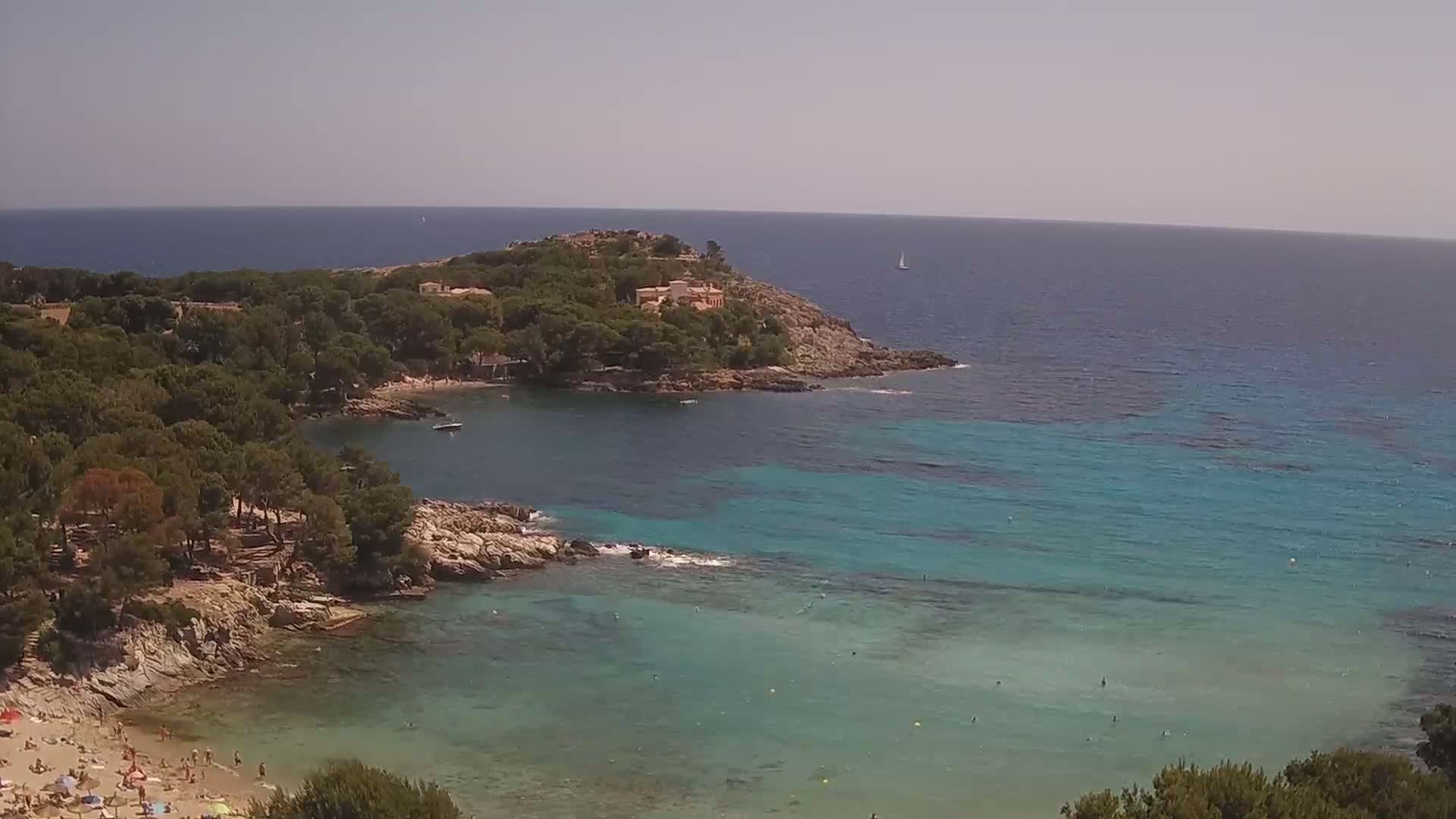 A secluded cove with a sandy beach and clear, turquoise water is surrounded by lush green vegetation, under a bright, sunny sky.