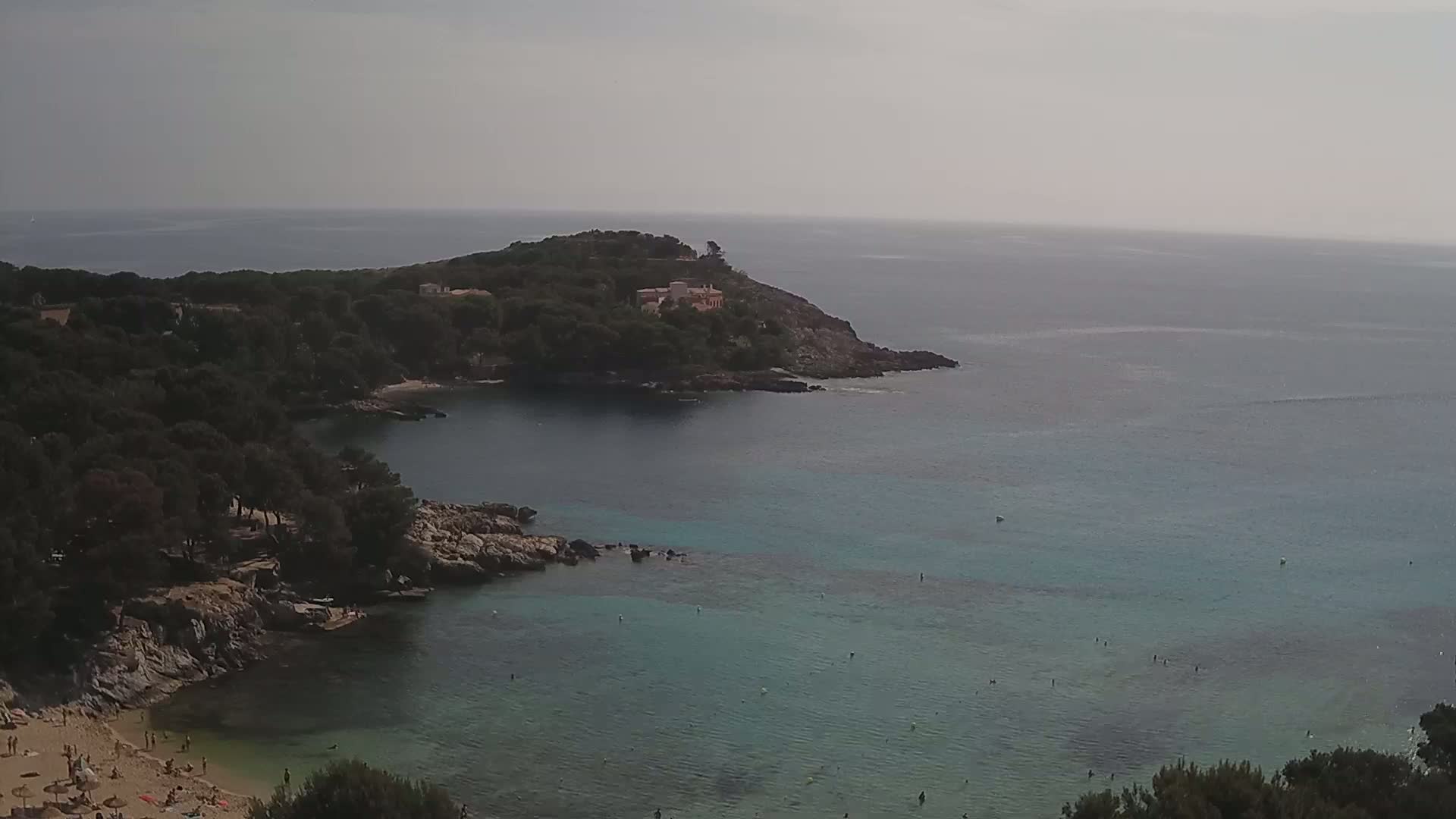 A secluded cove with a sandy beach and clear, shallow turquoise water is surrounded by dark green vegetation and rocky shoreline under an overcast sky.