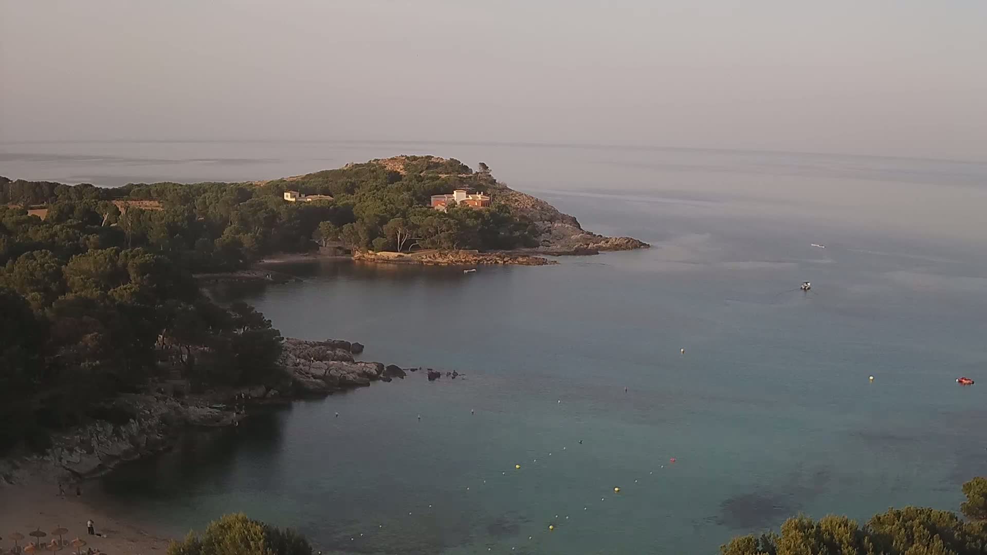 A tranquil, hazy coastal scene shows a small cove with a sandy beach, rocky shoreline, and calm, clear turquoise water, featuring a house perched atop a verdant hill.