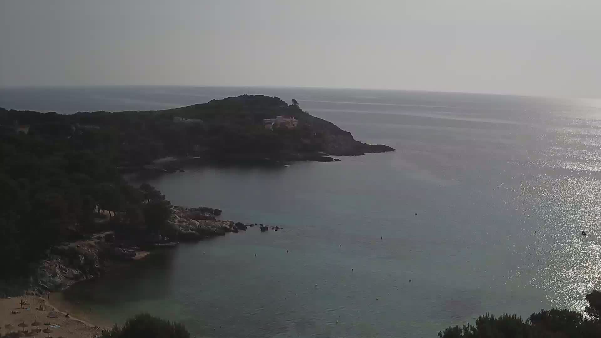 A calm, sunlit sea laps a secluded cove bordered by dark green vegetation and a small, sandy beach.