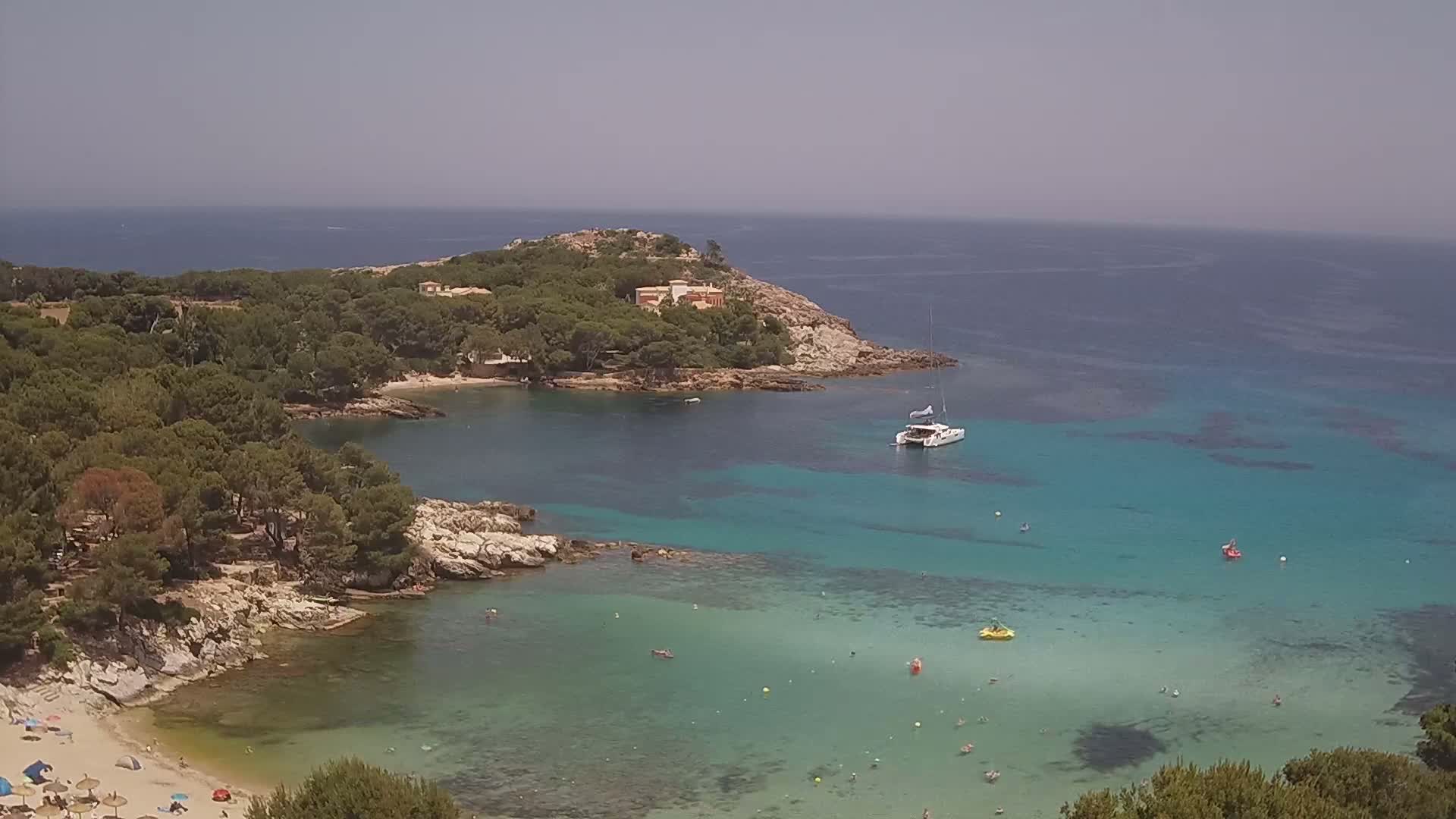 A secluded cove with clear turquoise water, a sandy beach, and a small building on a tree-covered headland under a clear sky.
