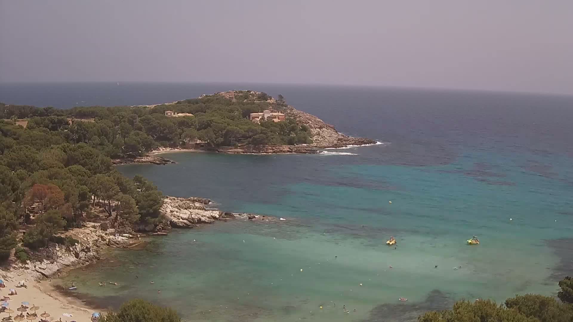 A secluded cove with a sandy beach and clear turquoise water is surrounded by pine trees and rocky outcrops under a hazy sky.