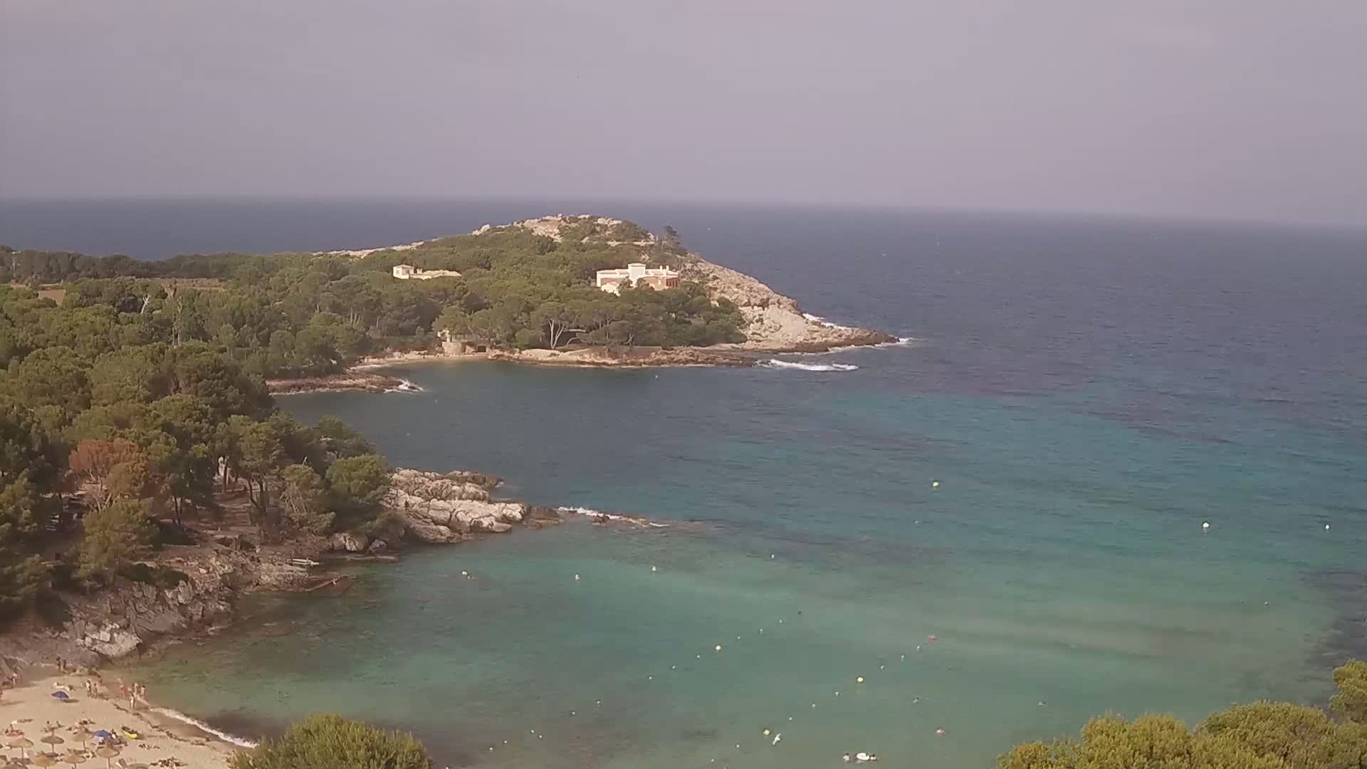 A tranquil cove with a sandy beach and clear turquoise water, nestled amongst pine-covered hills under an overcast sky.