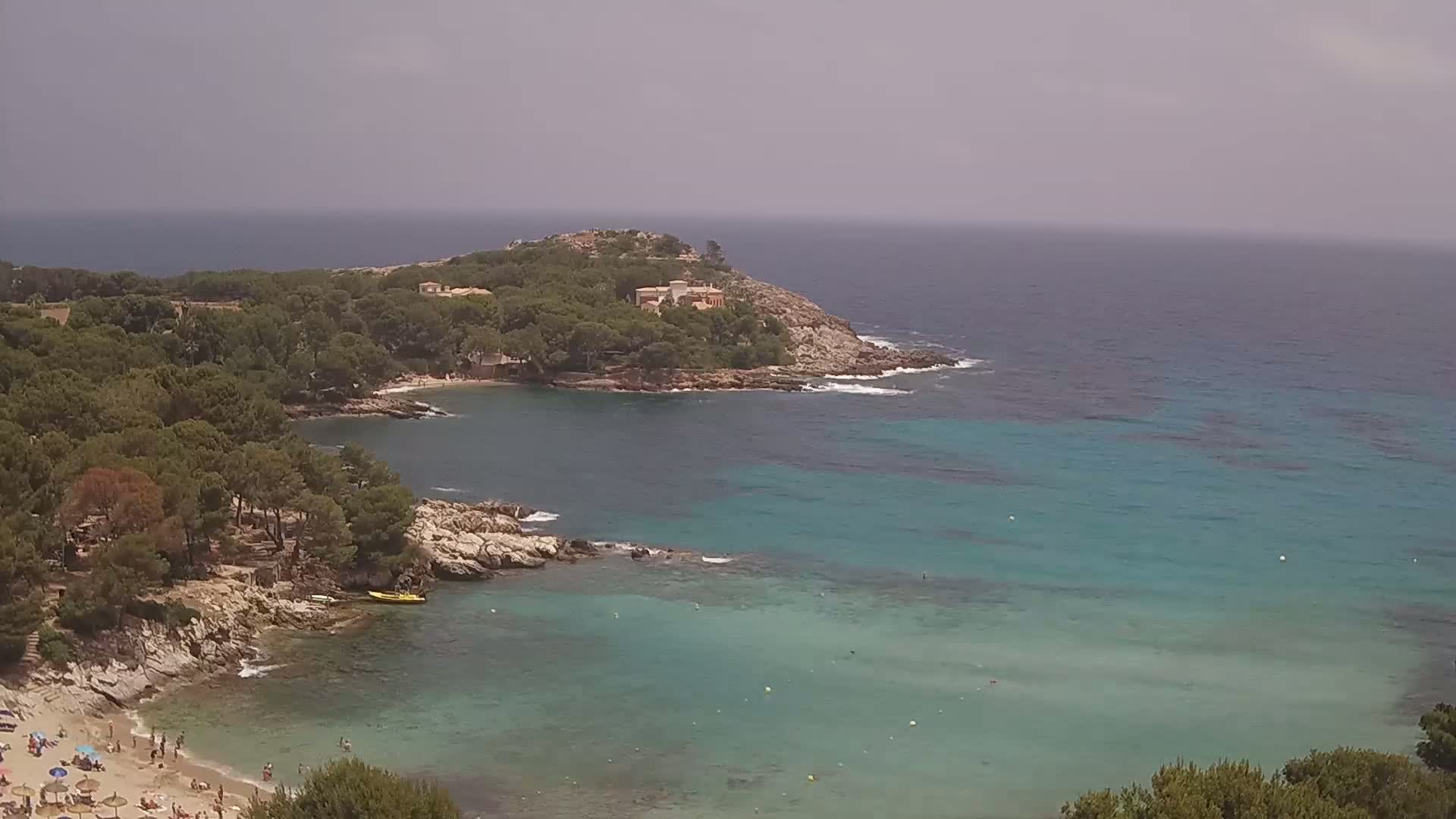 A secluded cove with a sandy beach and clear turquoise water, surrounded by pine-covered hills under an overcast sky.