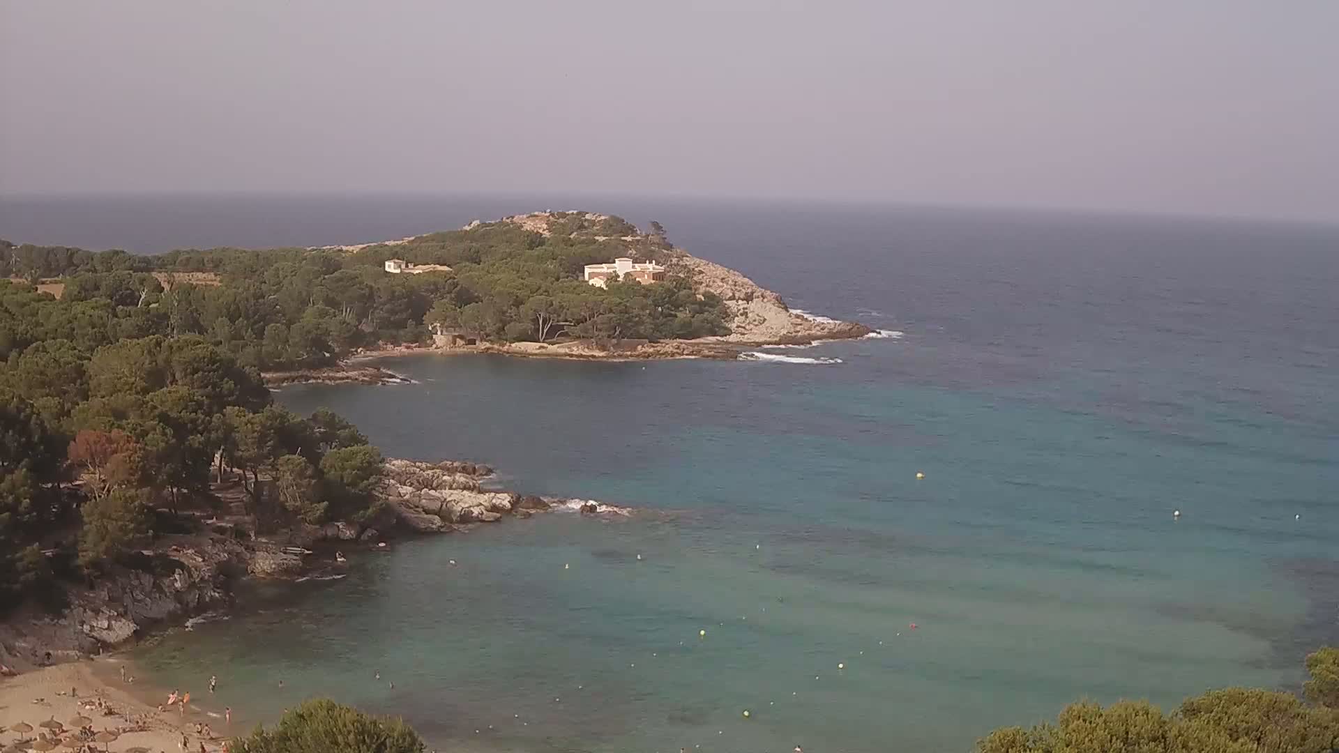 A secluded cove with a sandy beach and clear turquoise water, surrounded by pine trees and a small house on a rocky headland, under a hazy sky.