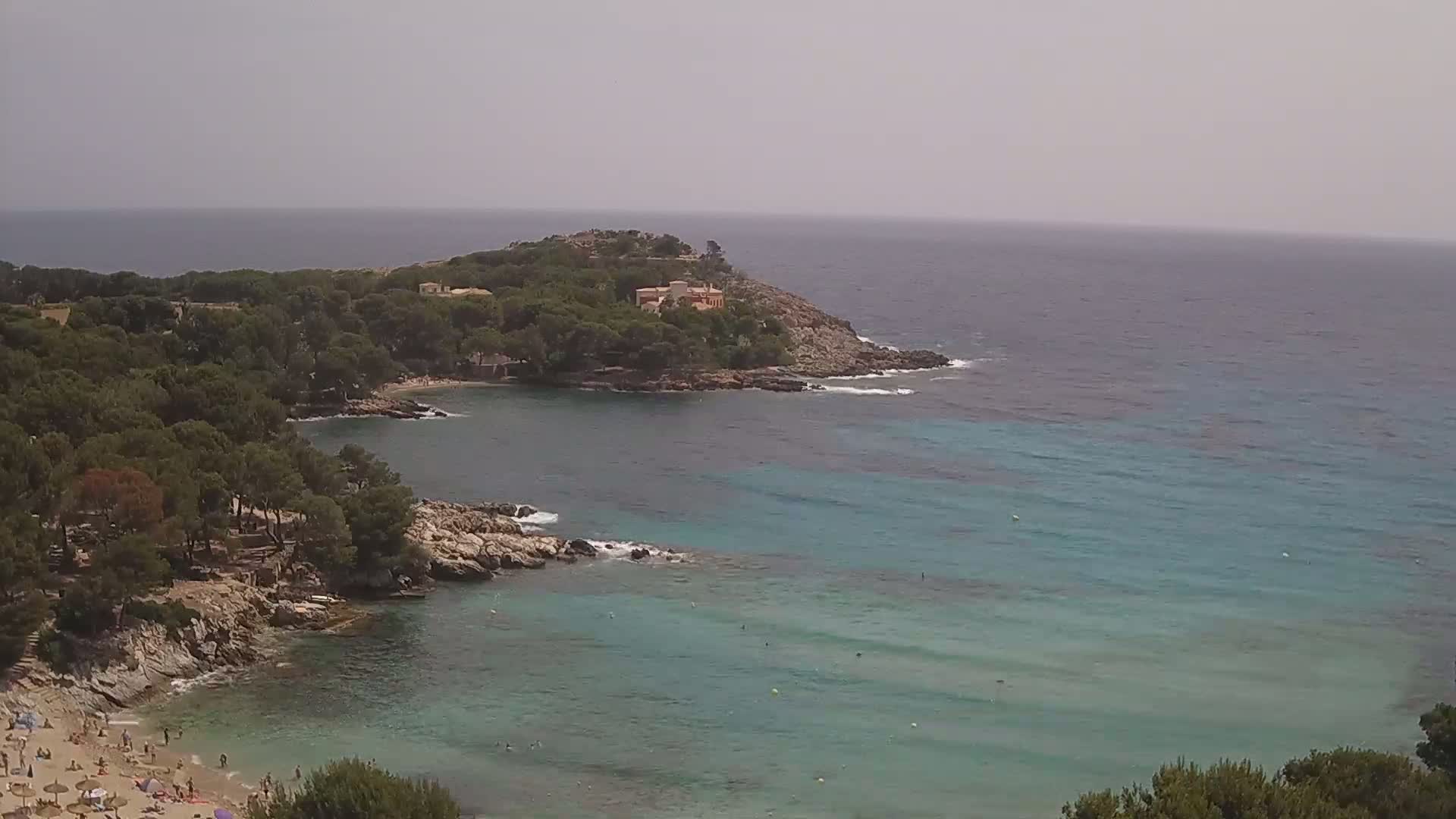 A secluded cove with a sandy beach and turquoise water is surrounded by lush green vegetation and rocky cliffs under an overcast sky.