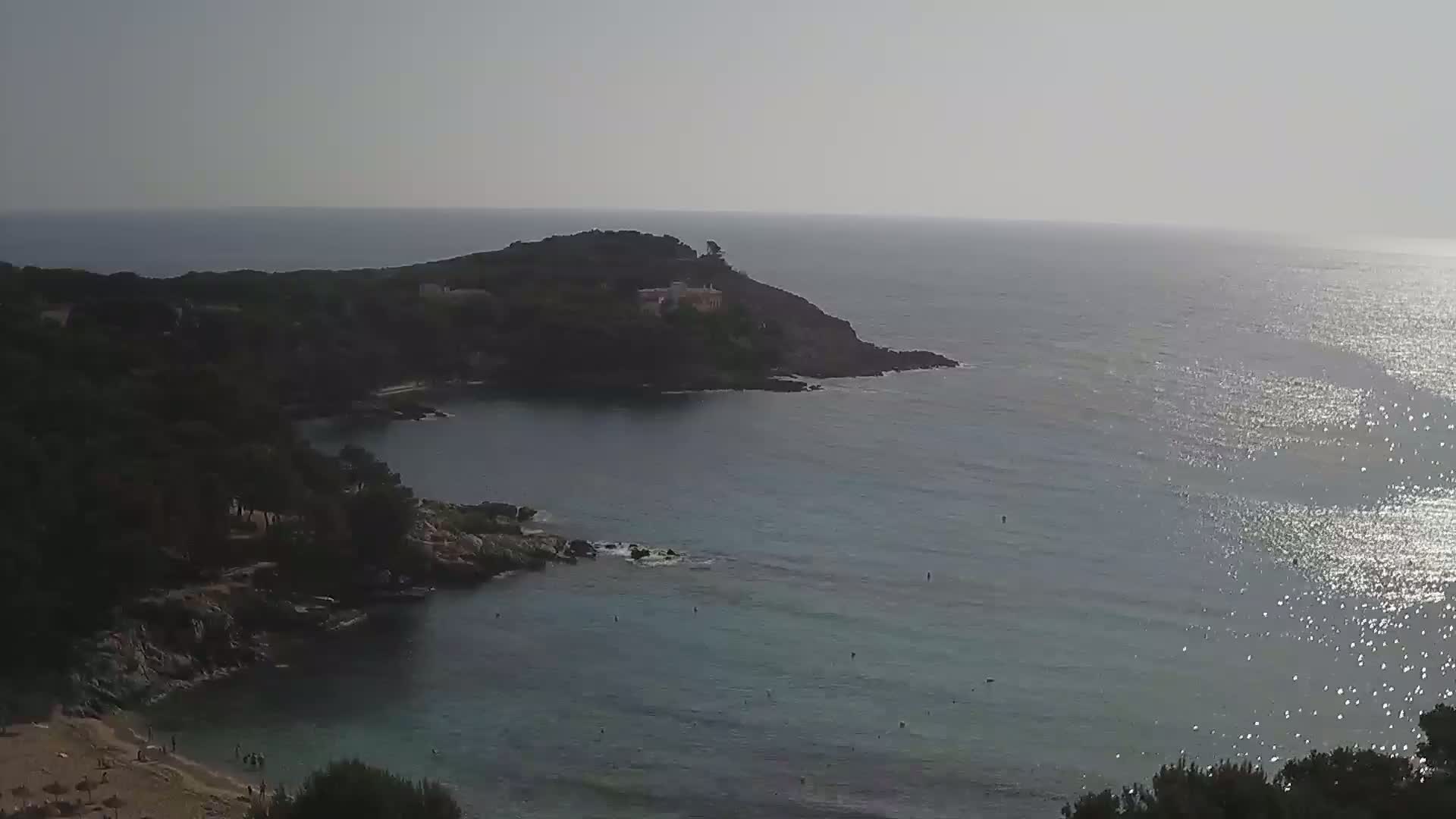 A calm, sunny day reveals a secluded cove with a small beach and a rocky headland extending into a glittering sea.