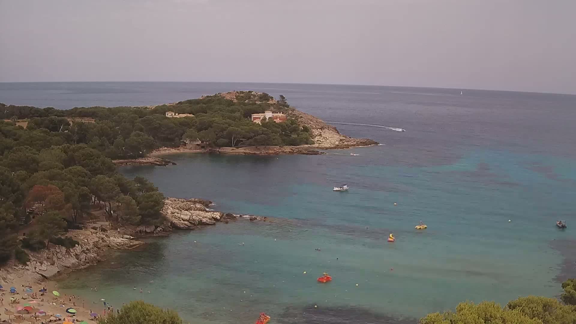 A secluded cove with turquoise water, a sandy beach with people, and a small island with buildings is visible under an overcast sky.