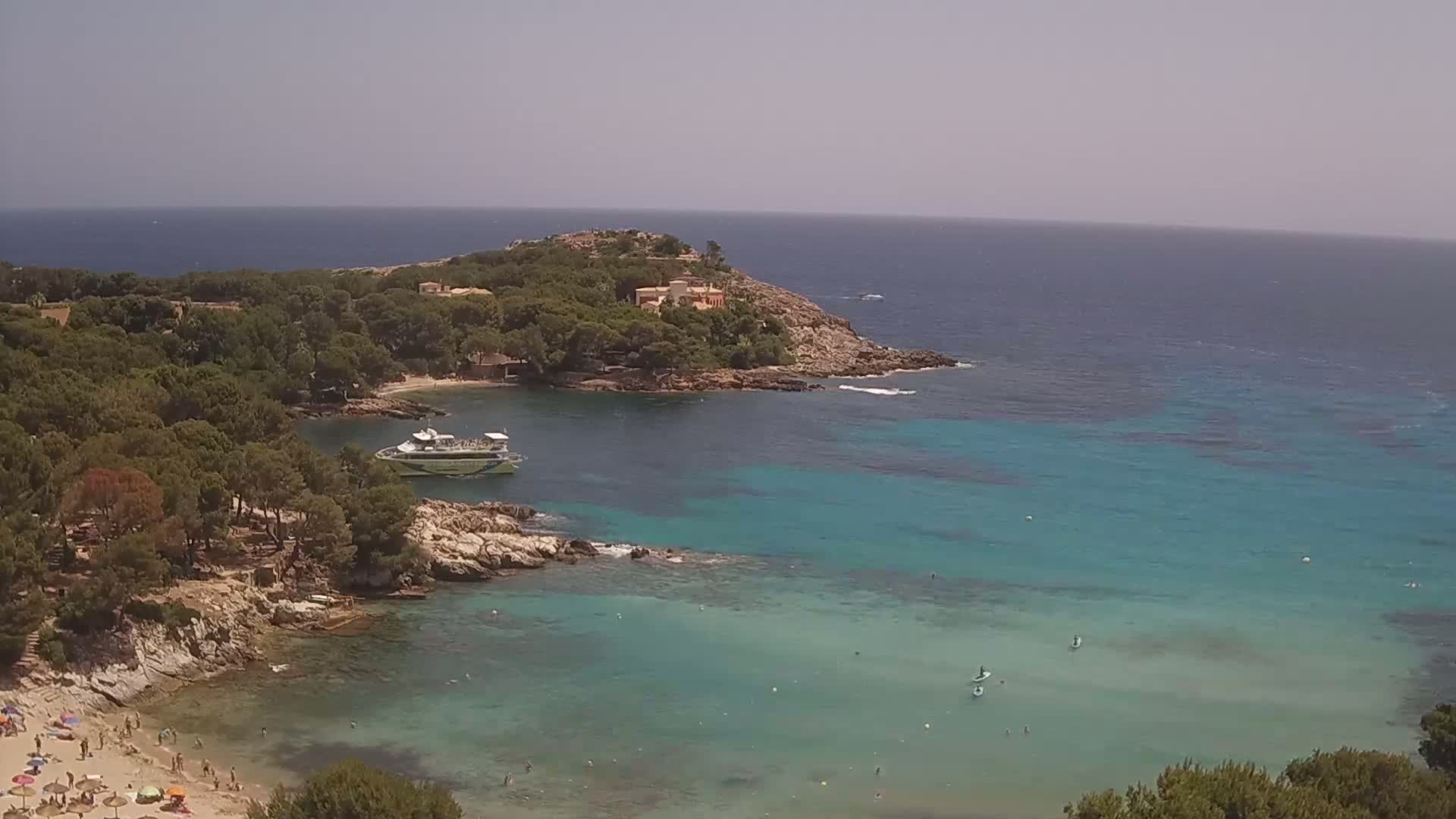 A tranquil, sunny day reveals a secluded cove with turquoise water, a boat, beachgoers, and a verdant headland.