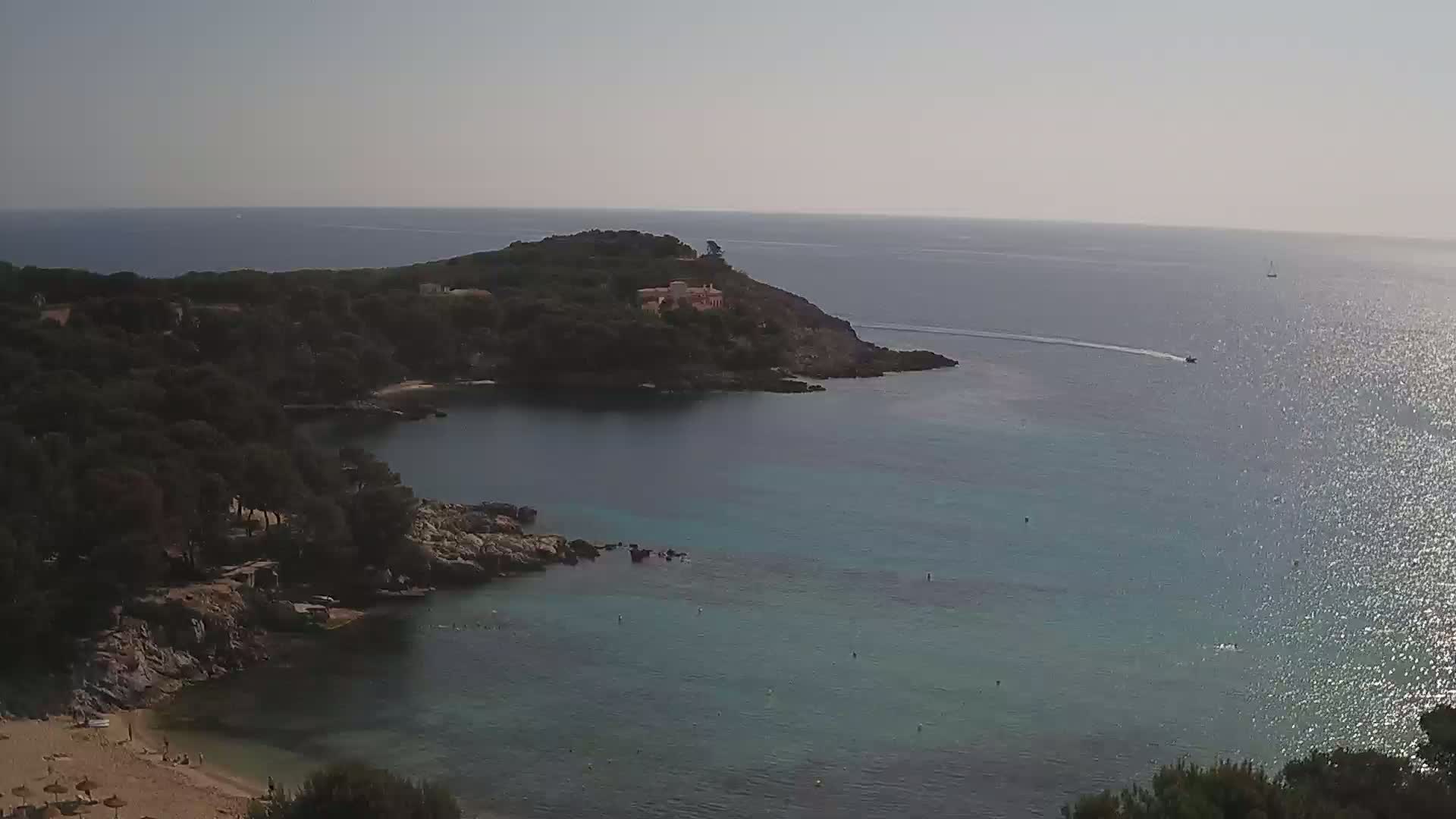 A sandy beach curves into a clear, calm bay surrounded by dark green trees and rocky shores, with a small building visible on a distant headland under a bright, sunny sky.