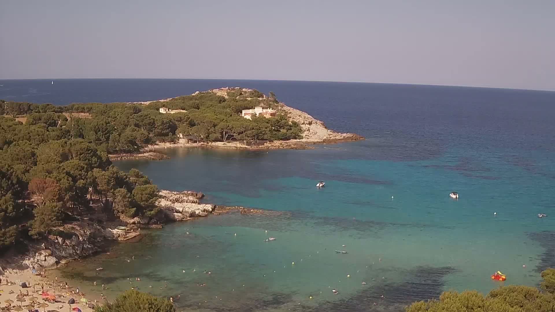 A secluded cove with clear turquoise water, a sandy beach with bathers, and surrounding pine trees under a sunny sky.